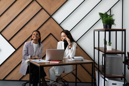 female colleagues in the office communicate while sitting at the table in front of a laptop.