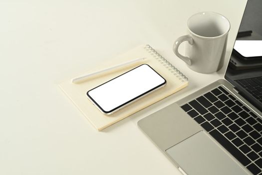 Mock up smart phone with white empty screen, notepad, coffee cup and laptop computer on white office desk.