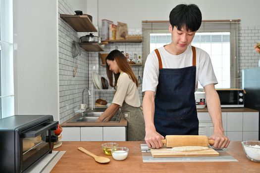 Smiling young asian man in apron using wooden roller pin making pastry dough in kitchen while preparing family dinner.