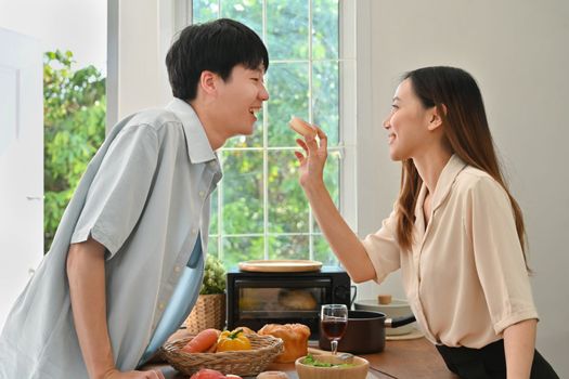 Lovely young woman feeding his boyfriends with donut while cooking in modern kitchen at home.