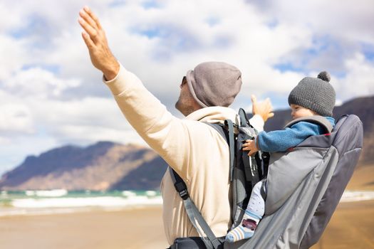 Young father rising hands to the sky while enjoying pure nature carrying his infant baby boy sun in backpack on windy sandy beach of Famara, Lanzarote island, Spain. Family travel concept