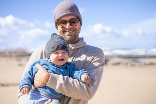 Father enjoying pure nature holding and playing with his infant baby boy sun in on windy sandy beach of Famara, Lanzarote island, Spain. Family travel and parenting concept