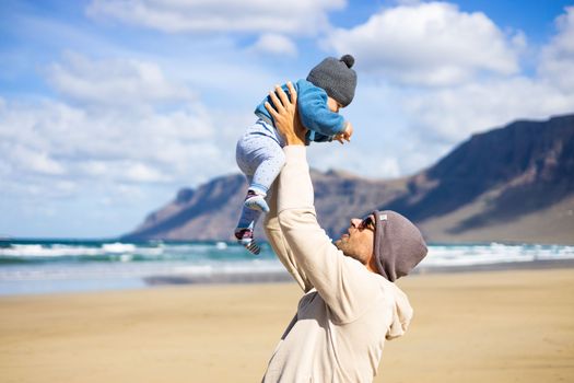 Father enjoying pure nature holding and playing with his infant baby boy sun in on windy sandy beach of Famara, Lanzarote island, Spain. Family travel and parenting concept