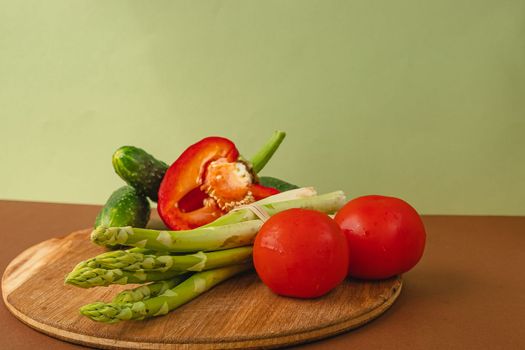 Vegetables lie on a wooden board: tomatoes, asparagus, cucumbers, red bell peppers. brown, light green background. place for text