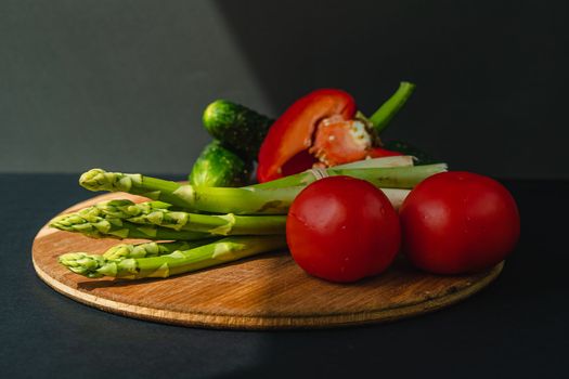 Vegetables lie on a wooden board: tomatoes, asparagus, cucumbers, red bell peppers. brown, dark gray background. place for text