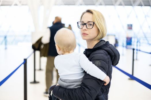 Mother holding his infant baby boy child queuing at airport terminal in passport control line at immigrations departure before moving to boarding gates to board an airplane. Travel with baby concept.