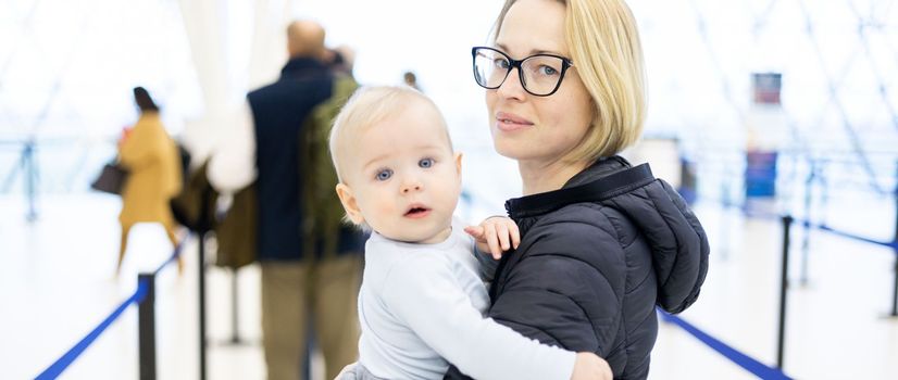 Mother holding his infant baby boy child queuing at airport terminal in passport control line at immigrations departure before moving to boarding gates to board an airplane. Travel with baby concept.