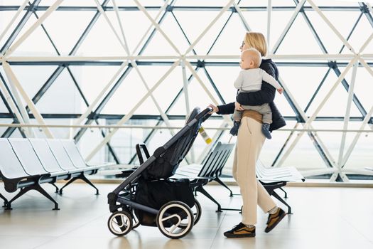 Mother carying his infant baby boy child, pushing stroller at airport departure terminal moving to boarding gates to board an airplane. Family travel with baby concept