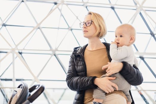 Mother carying his infant baby boy child, pushing stroller at airport departure terminal waiting at boarding gates to board an airplane. Family travel with baby concept