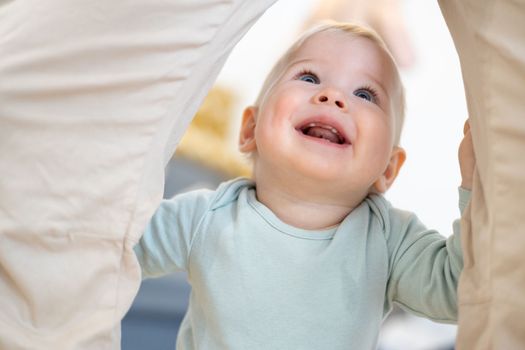 Portrait of adorable cheerful infant baby boy child taking first steps holding to father's pants at home. Cute baby boy learning to walk