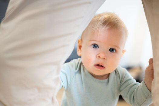 Portrait of adorable curious infant baby boy child taking first steps holding to father's pants at home. Cute baby boy learning to walk