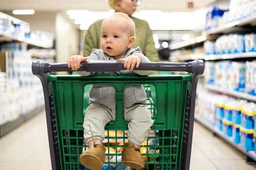 Mother pushing shopping cart with her infant baby boy child down department aisle in supermarket grocery store. Shopping with kids concept
