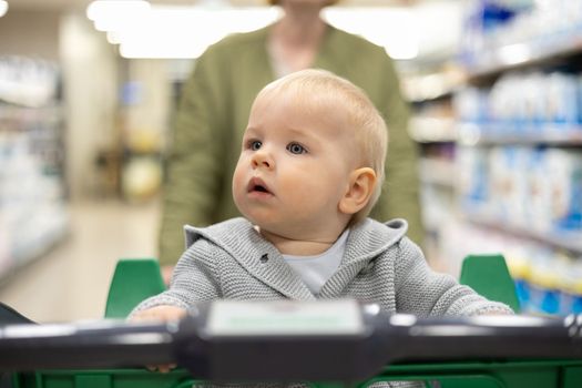 Mother pushing shopping cart with her infant baby boy child down department aisle in supermarket grocery store. Shopping with kids concept