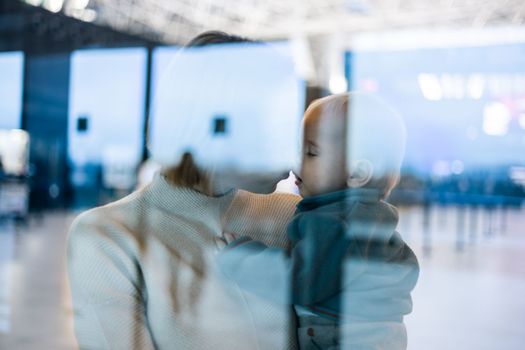 Thoughtful young mother kissing and caressing his infant baby boy child while waiting by the window at airport terminal departure gates. Travel with baby concept