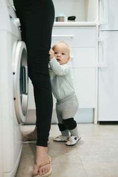 Little infant baby boy child hiding between mothers legs demanding her attention while she is multitasking, trying to do some household chores in kitchen at home. Mother on maternity leave