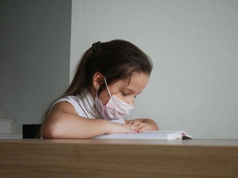 Distance learning online education. Sickness schoolgirl in medical mask studying at home with book doing school homework. Training book on table