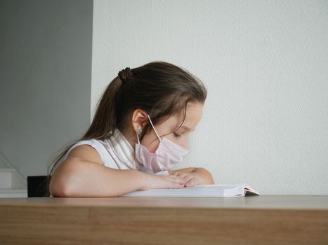 Distance learning online education. Sickness schoolgirl in medical mask studying at home with book doing school homework. Training book on table