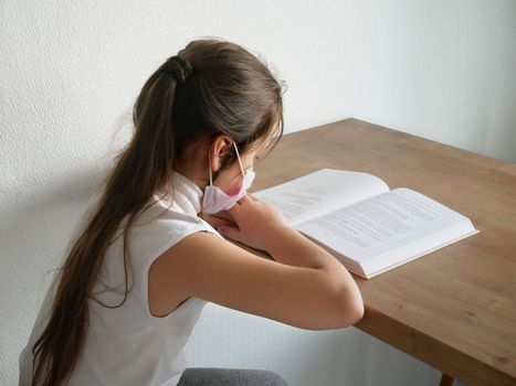 Distance learning online education. Sickness schoolgirl in medical mask studying at home with book doing school homework. Training book on table