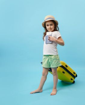 Full length portrait of cute little traveler girl with yellow suitcase, showing flight ticket or boarding pass to camera, isolated on blue colored background. Kid going for summer vacations. ad space
