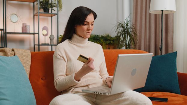 Young woman using credit bank card and laptop computer while transferring money, purchases online shopping, order food delivery at home apartment. Adult girl in day living room sitting on couch indoor