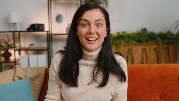 Oh my God, Wow. Surprised woman with brunette hair at home couch, shocked by sudden victory triumph, good win news, celebrating lottery jackpot luck. Excited amazed girl in living room apartment