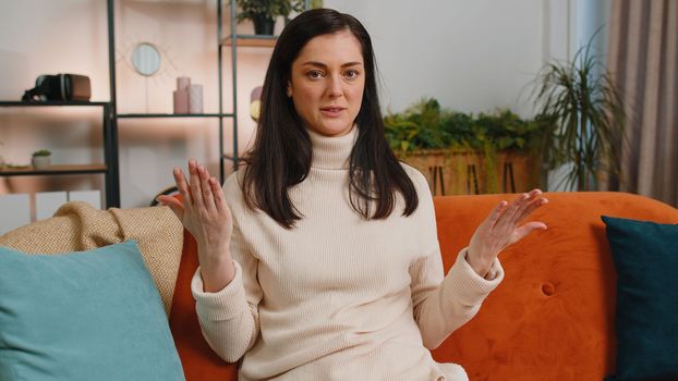 What. Why. Young woman raising hands in indignant expression asking reason of failure demonstrating disbelief irritation by troubles at home apartment. Brunette girl in living room sitting on sofa