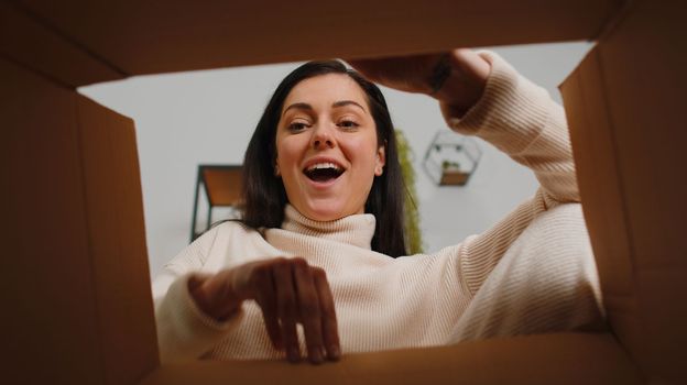 Happy surprised winner young woman unpacking delivery parcel at home. Smiling satisfied girl shopper online shop customer opening cardboard box receiving purchase gift present by fast postal shipping