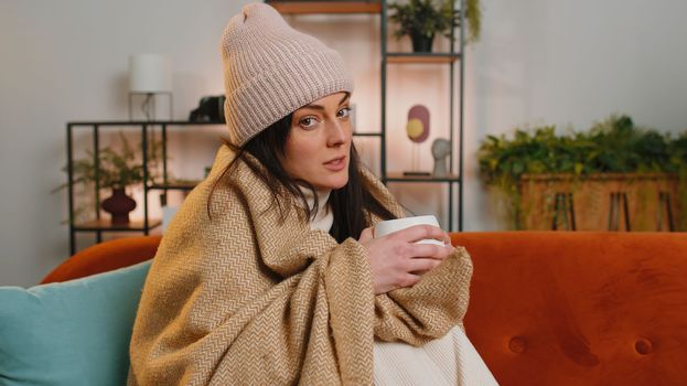 Sick woman wrapped in plaid sits alone shivering from cold on sofa drinking hot tea in unheated apartment without heating due debt. Unhealthy ill girl in hat feeling discomfort try to warm up at home