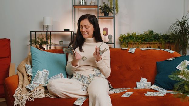 Successful happy rich business woman throwing money cheering up with high profits, lottery game win, satisfied of wealth income earnings salary. Girl at home room couch full of dollar cash banknotes