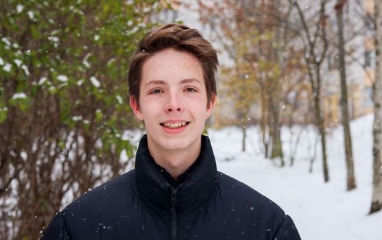 Portrait of a teenage boy in front of an apartment building and a bush in winter. The boy poses on a snowy street and looks at the camera.
