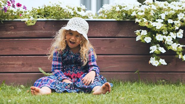 A little girl in a panama hat sits by the white petunias in the garden
