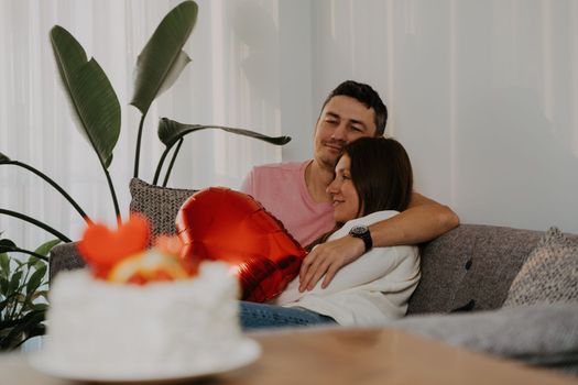 Beautiful young couple sitting on a couch, hugging on romantic date on Valentine day at home. Wife and husband celebrate with heart shape ballon, cake, flowers.