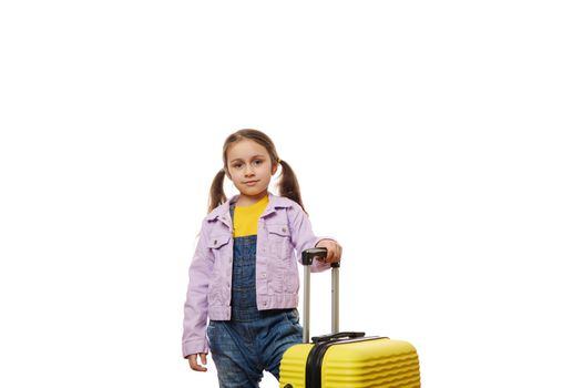 Caucasian cute baby girl, little traveler child dressed in casual denim overalls and purle jacket, standing near her yellow suitcase isolated over white background. Tourism. Travel. Advertising space