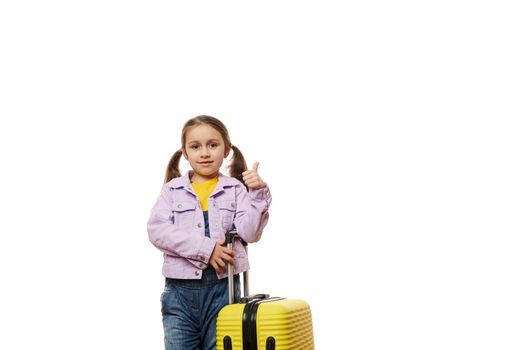 Adorable traveler child, lovely baby girl in violet denim jacket and blue overalls, going for vacations, showing thumb up looking at camera, posing with yellow suitcase over white background