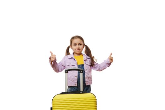 Adorable baby girl with two ponytails, dressed in purple jacket and blue denim overalls, standing behind a yellow suitcase, demonstrating thumb up looking at camera, isolated over white background