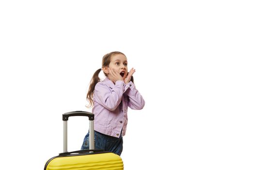 Amazed little traveler girl dressed in purple jacket and blue denim overalls, with surprised look, expressing wow emotion, standing with yellow luggage on white background with free advertising space
