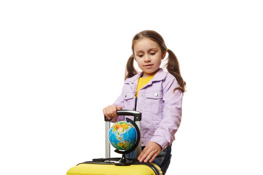 Beautiful Caucasian little child traveler tourist, lovely baby girl with two ponytails, in purple denim jacket, posing with globe and yellow suitcase on white background. Worldwide tourism concept