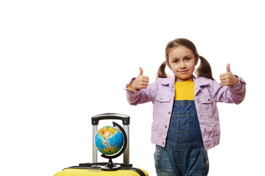 Adorable delightful baby girl wearing purple denim jacket and blue overalls showing thumbs up at camera, standing next to a globe on her yellow suitcase, isolated on white background. Copy ad space