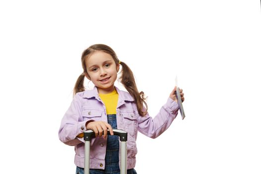 Adorable Caucasian little traveler girl in purple jacket, going for vacations, holding suitcase and boarding pass, cutely smiling looking at camera, isolated on white background with copy ad space