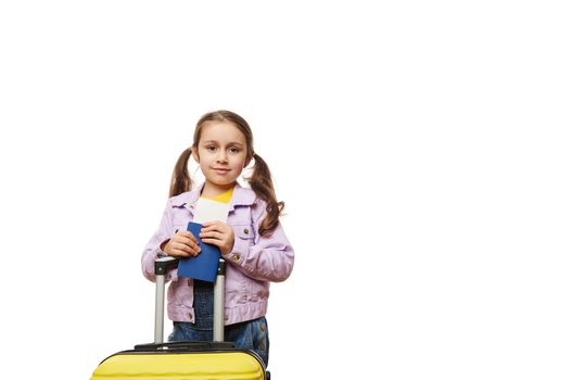 Beautiful 5 years old child, Caucasian lovely little traveler girl with yellow suitcase, going for summer vacation, holding boarding pass flight ticket, isolated on white background. Advertising space