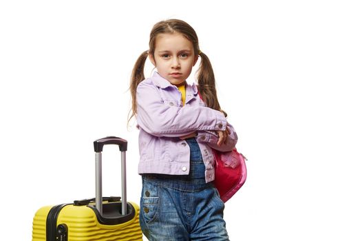 Isolated portrait on white background of a little child girl with two ponytails, dressed in purple jacket, posing with her arms crossed, standing near yellow suitcase over white background. Copy Space