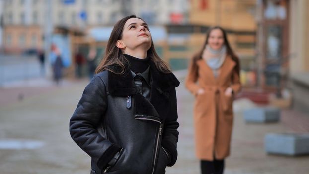 A friend comes up from behind her waiting girlfriend on a city street