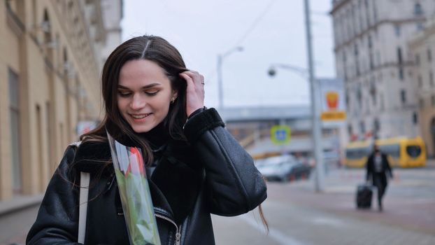 A happy brunette girl in a black jacket walks through the city in early spring