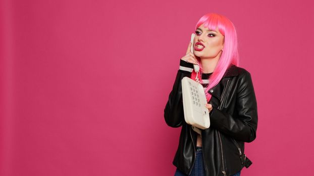 Happy confident model with pink hair talking on landline phone call, chatting on retro telephone line with cord in studio. Having fun using vintage phone for remote communication, cool person.