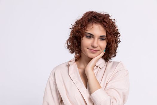 A young girl gently touches her cheek with her hand. Beautiful curly chestnut-colored hair. Natural hairstyle. Light-colored long-sleeved blouse.