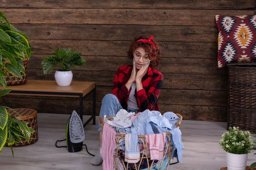 The young woman does not believe that for today she has so many men's shirts to iron. Hands laid on cheeks in surprise. A wall in a house made of boards.
