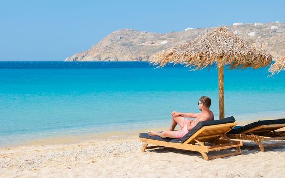 Young men on the beach of Mykonos beach during summer with umbrella and luxury beach chairs beds, blue ocean with the mountain at Elia beach Mikonos Greece.