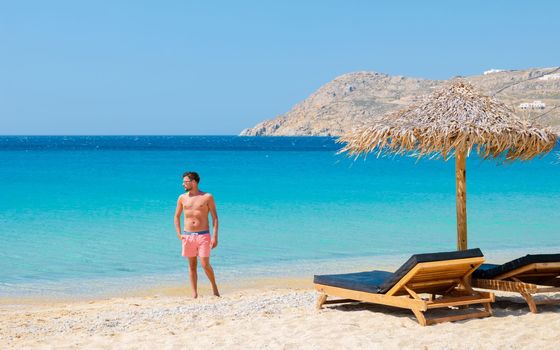 Young men on the beach of Mykonos beach during summer with umbrella and luxury beach chairs beds, blue ocean with the mountain at Elia beach Mikonos Greece.