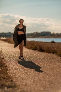 Portrait blonde sea cape. A calm young blonde in an unbuttoned khaki raincoat walks along the seashore, under a raincoat a black skirt and top.