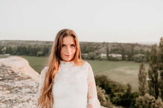 Romantic beautiful bride in white dress posing with sea and mountains in background. Stylish bride standing back on beautiful landscape of sea and mountains on sunset